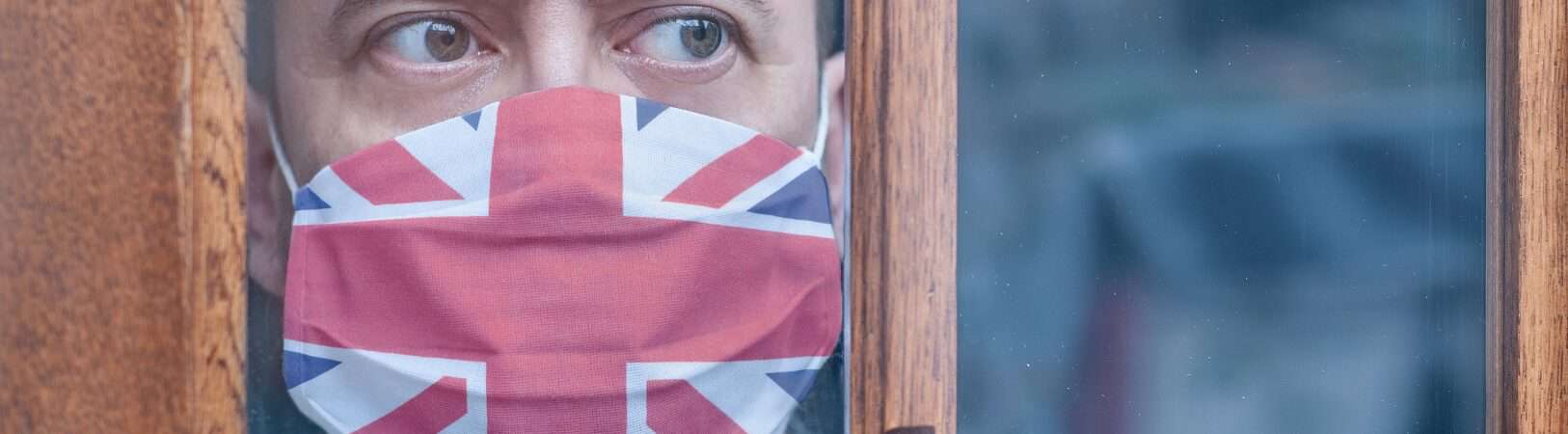 Jovem com uma máscara protetora com uma bandeira britânica olhando pela janela