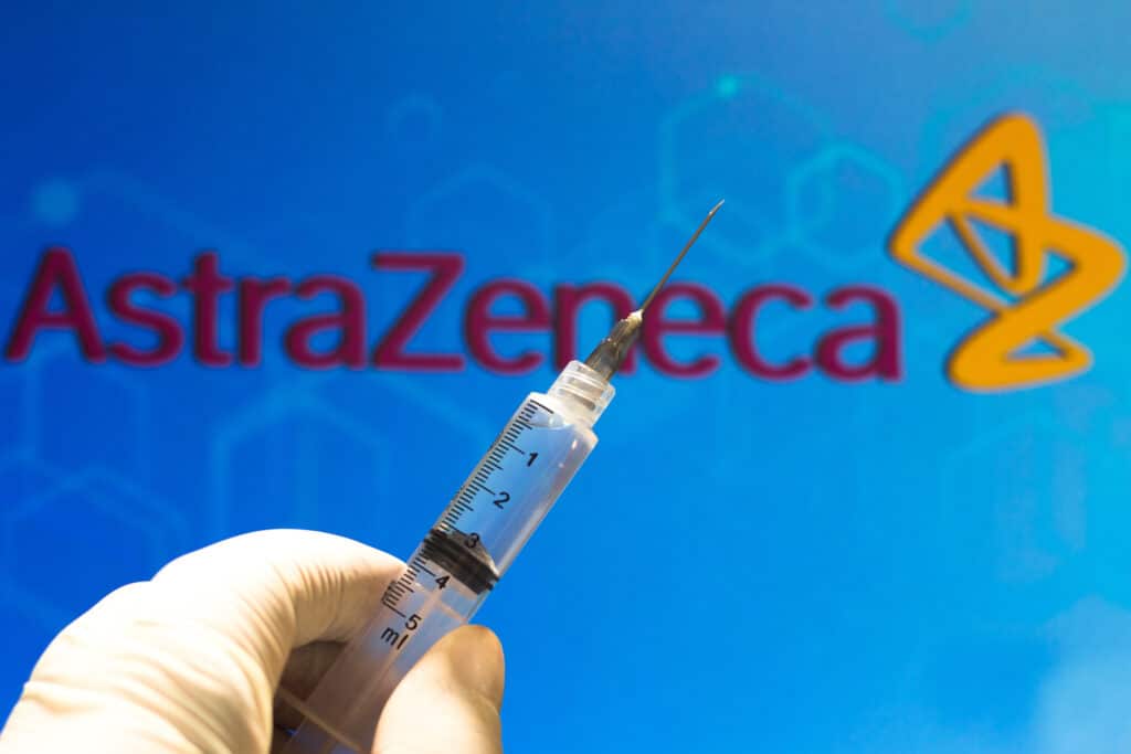 Mão com luva segura seringa com agulha em frente a logomarca da farmacêutica AstraZeneca