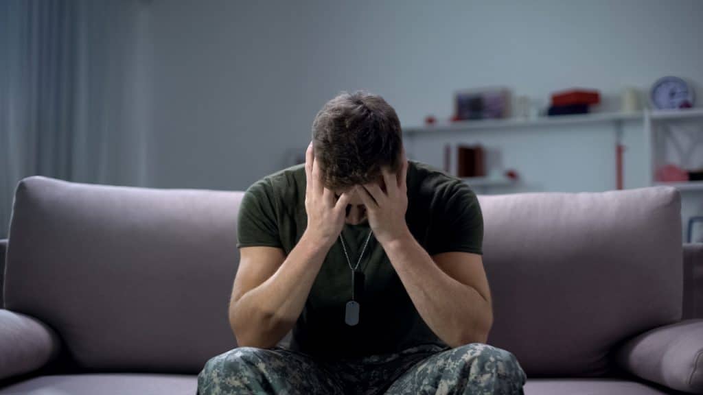 Imagem mostra um soldado sentado em um sofá, com as mãos segurando o rosto em tom de tristeza, simbolizando a depressão