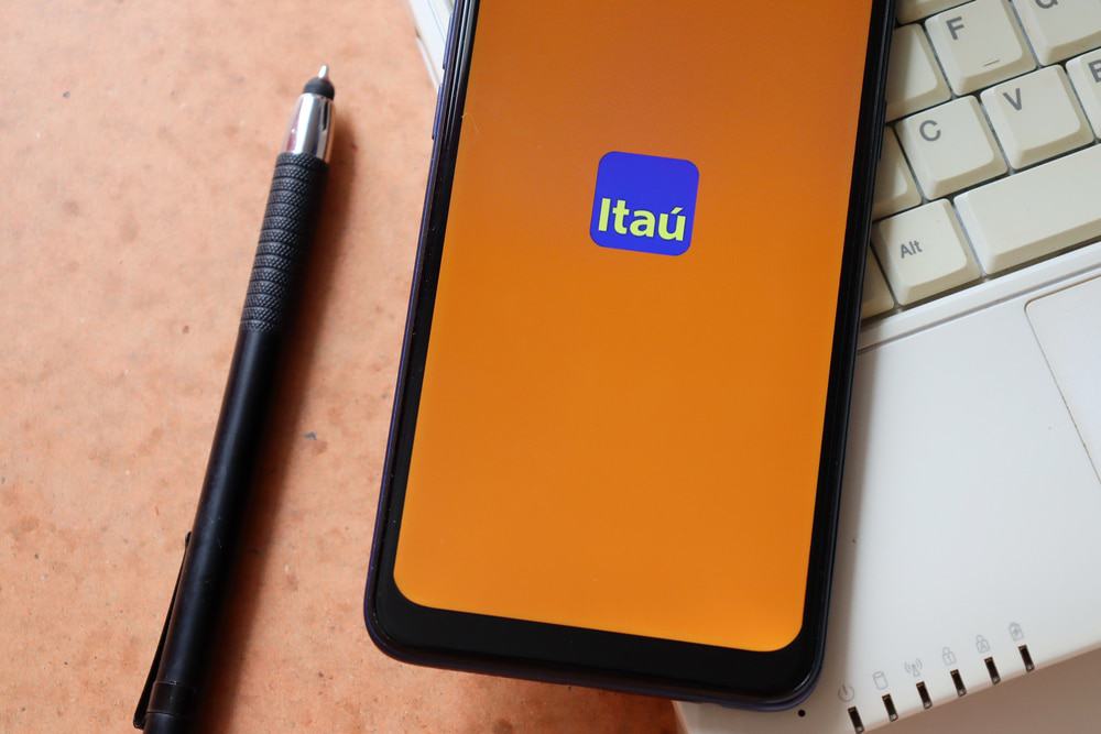 Logotipo do banco Itaú aparece em tela de smartphone, que por sua vez está em cima de um teclado de notebook e ao lado de uma caneta.