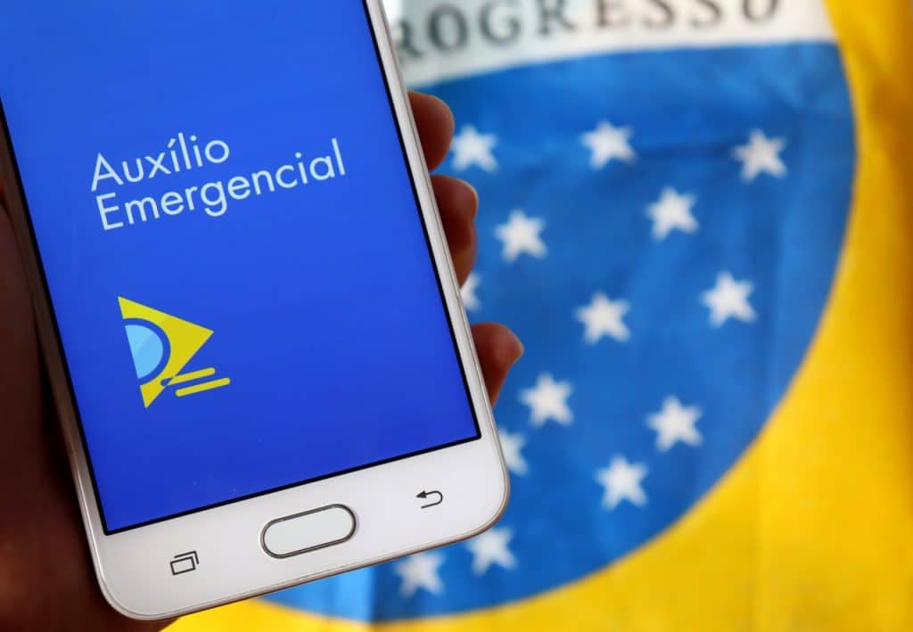 Smartphone exibindo o app do auxílio emergencial com uma bandeira do Brasil ao fundo