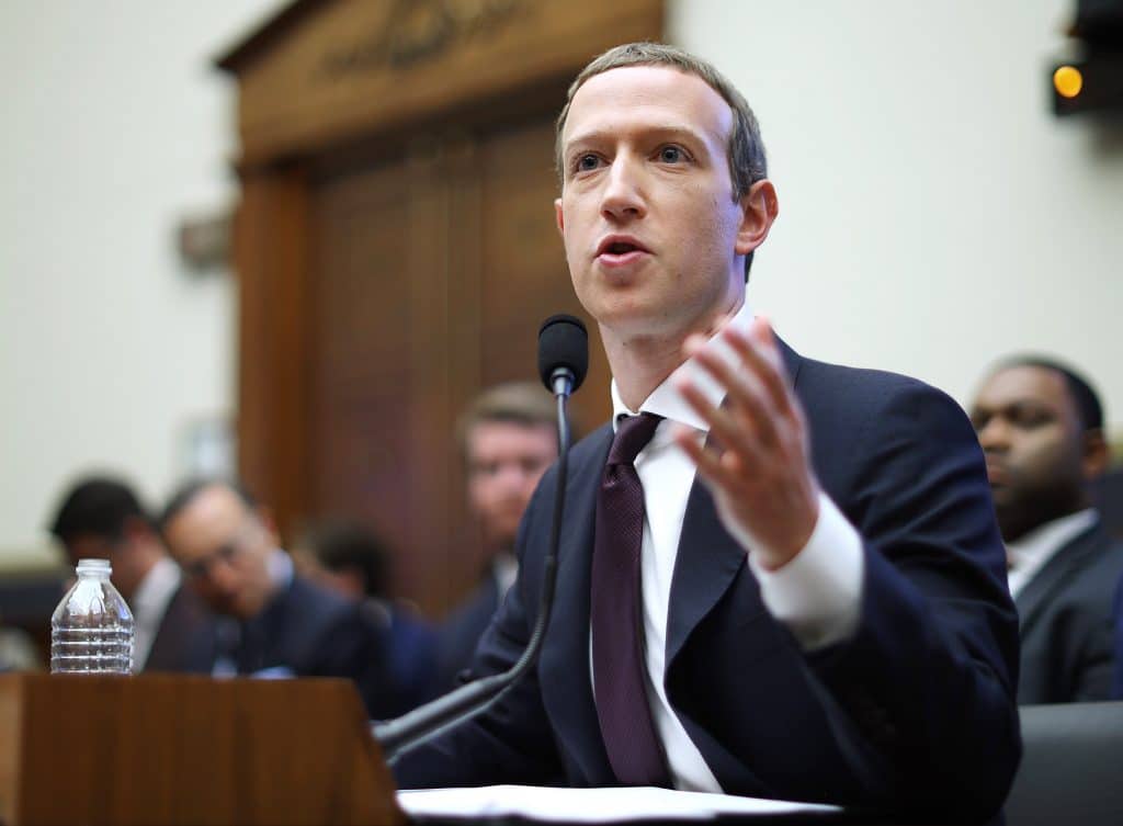 Inagem mostra o CEO do Facebook, Mark Zuckerberg, sentado em um acadeira de madeira, respondendo a perguntas durante uma audiência. Ele veste terno preto com gravata da mesma core está com o cabelo curto