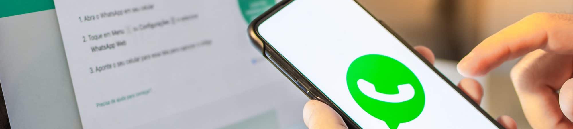 Imagem mostra uma mão segurando um smartphone com o logotipo do WhatsApp na tela; ao fundo há um computador, para demonstrar o uso do WhatsApp Web