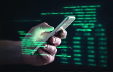 Imagem mostra uma mão segurando um smartphone e, ao redor dela, uma série de códigos e dados em fonte verde, simbolizando o acesso a informações privadas e golpes cibernéticos
