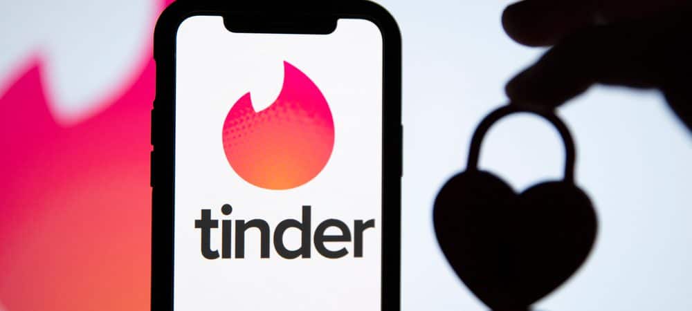 Ao fundo é possível ver a logomarca do Tinder, à frente aparece uma mão segurando um cadeado em formato de coração à direita; à esquerda está um smartphone com o logo do Tinder na tela