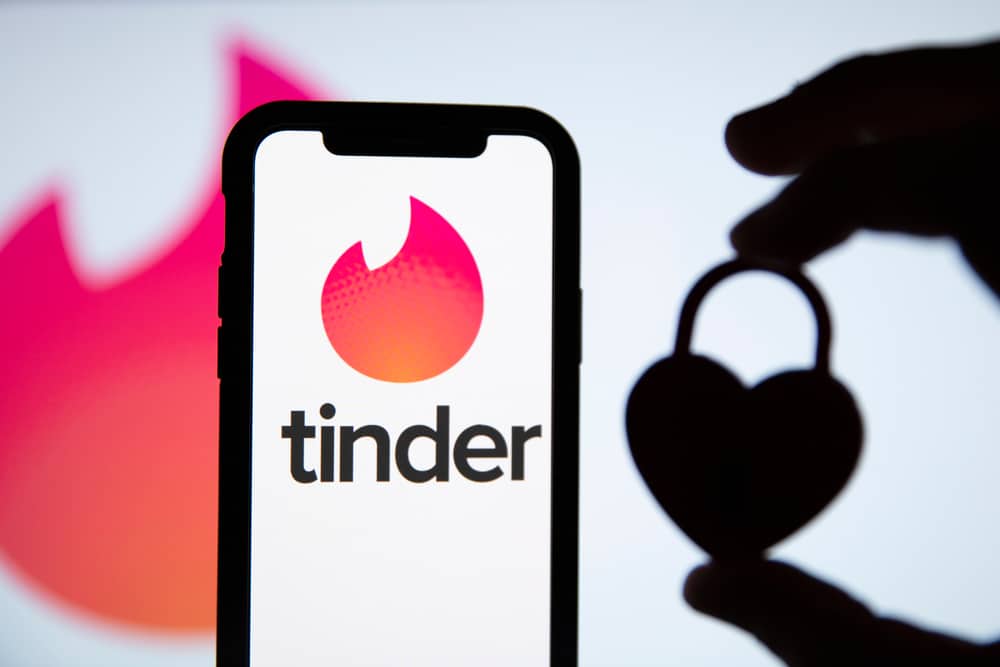 Ao fundo é possível ver a logomarca do Tinder, à frente aparece uma mão segurando um cadeado em formato de coração à direita; à esquerda está um smartphone com o logo do Tinder na tela