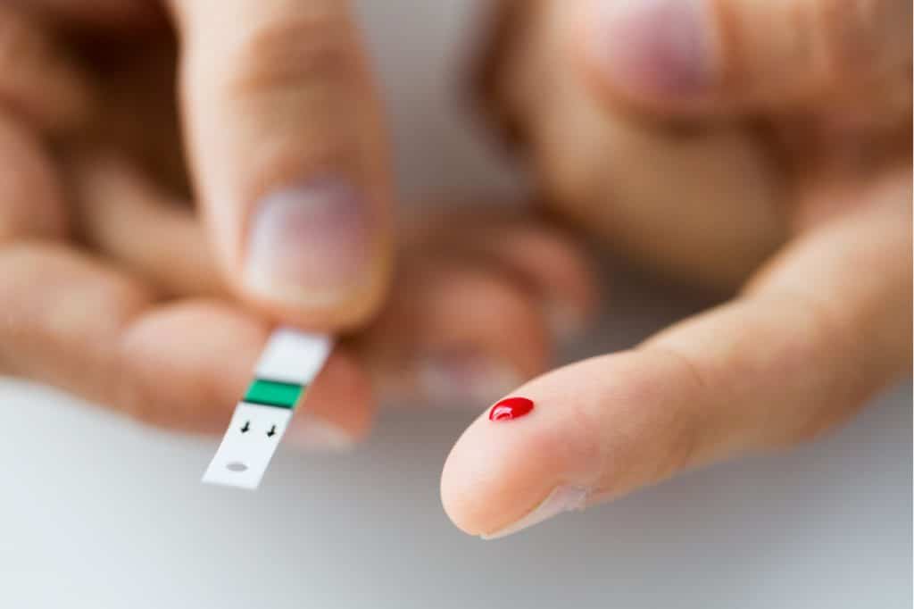 Imagem mostra um dedo perfurado, com uma gota de sangue à mostra. Na outra mão, um teste de diabetes