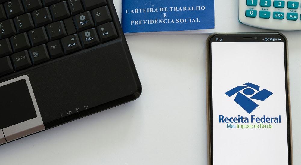 Computador, carteira de trabalho e celular mostrando tela da Restituição do Imposto de Renda