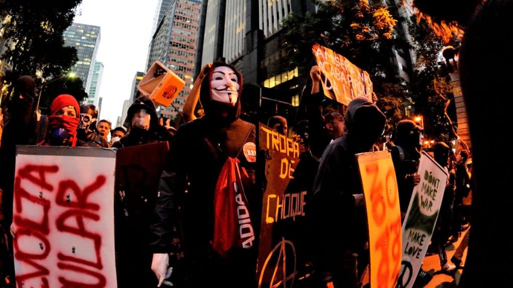 manifestante usa máscara do anonymous junto a bandeira antifa e escudo escrito tropa de choque com símbolo anarquista 