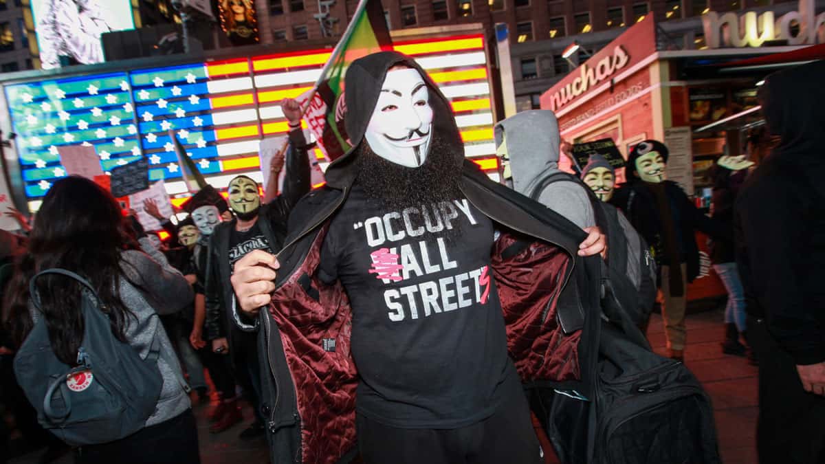 manifestante com máscara usando camiseta escrito occupy all streets em protesto nos estados unidos junto a diversas pessoas mascaradas