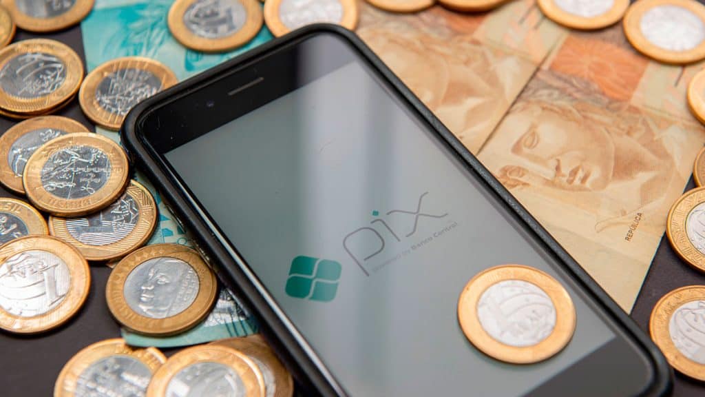 Celular com app do pix ao lado de duas notas de 50 reais e várias moedas de 1 real