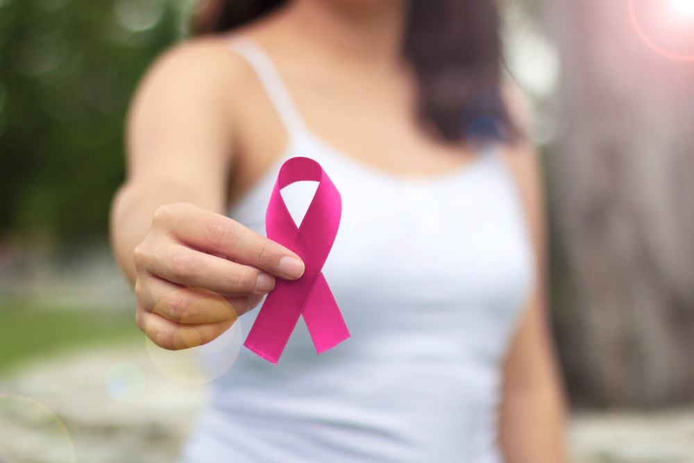 Imagem mostra o torso de uma mulher ao fundo, à frente ela segura um laço rosa, símbolo da campanha Outubro Rosa, de prevenção ao câncer de mama