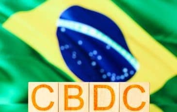 Cubos de madeira com a inscrição CBDC (Real Digital) com a bandeira do Brasil ao fundo