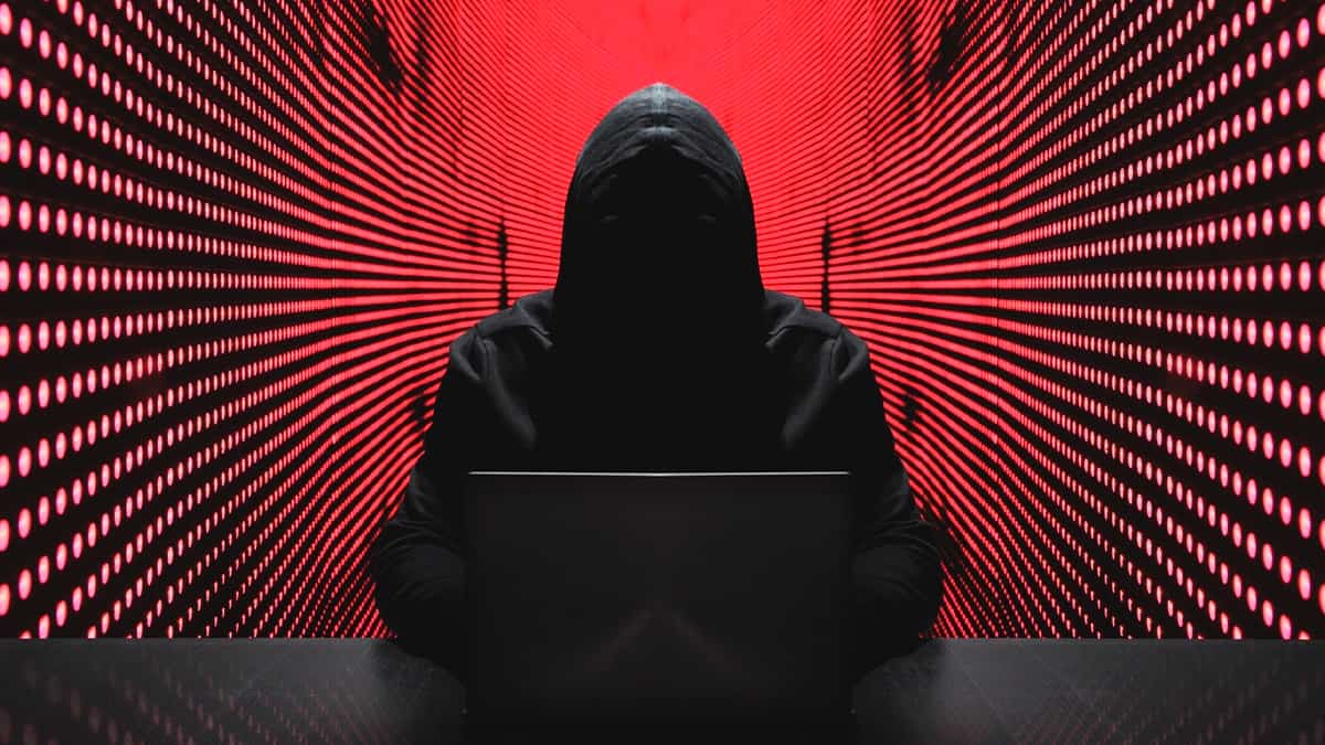 Hacker encapuzado sentado usando notebook numa mesa e, ao fundo, paredes com luzes vermelhas