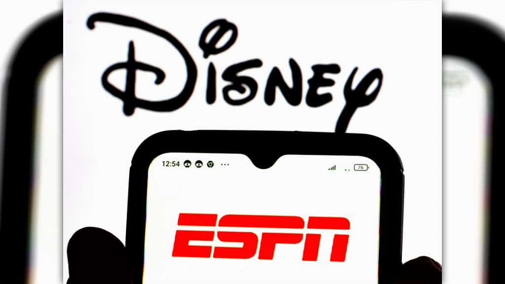 Pessoa segurando celular com logomarca da ESPN e, ao fundo, logomarca da Disney está aberta na tela de um computador