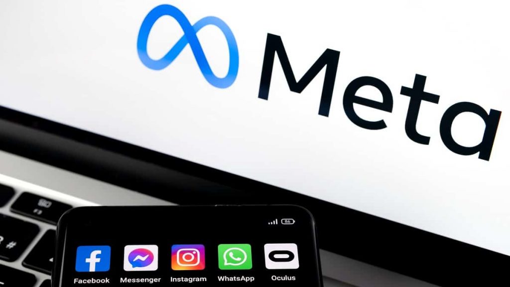 Celular com ícones de redes sociais da Meta e, ao fundo, o logotipo da marca aparece numa tela de computador